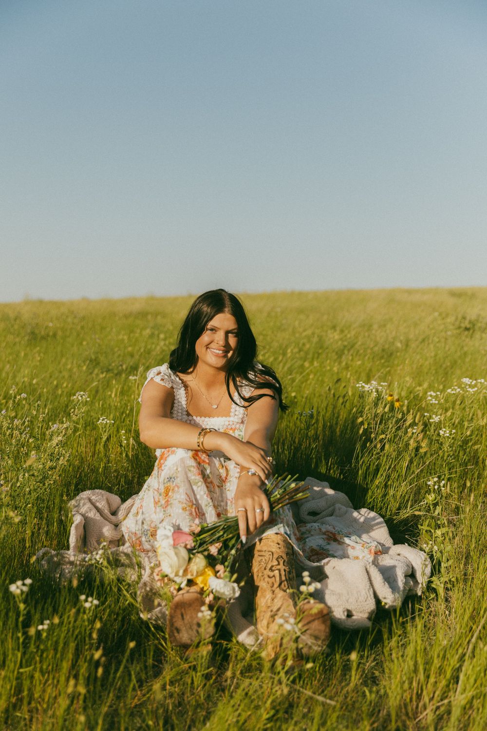 A person in a floral dress sits in a sunny meadow with tall grass and wildflowers on a warm summer day.