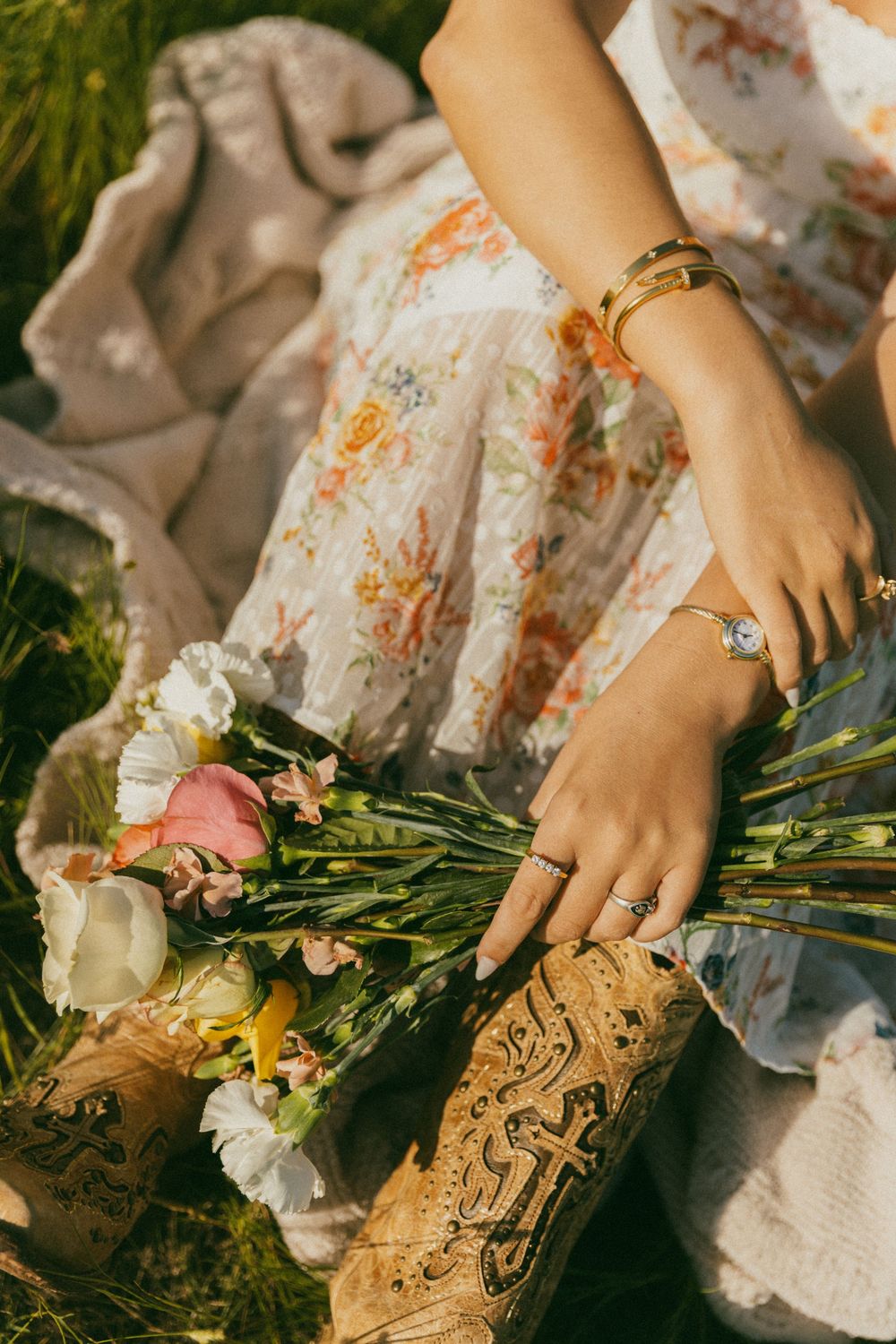 Hands adorned with jewelry hold a bouquet of fresh flowers against a floral dress and rustic cork background.