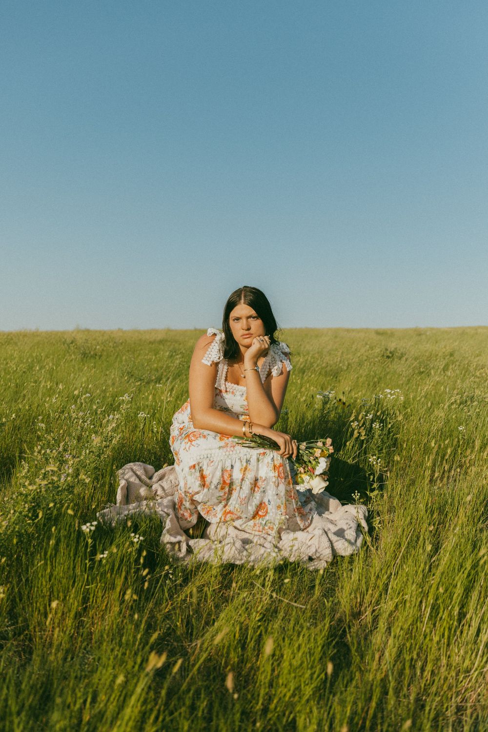 A person in a floral dress lounges gracefully in a sunlit meadow of tall grass and wildflowers.