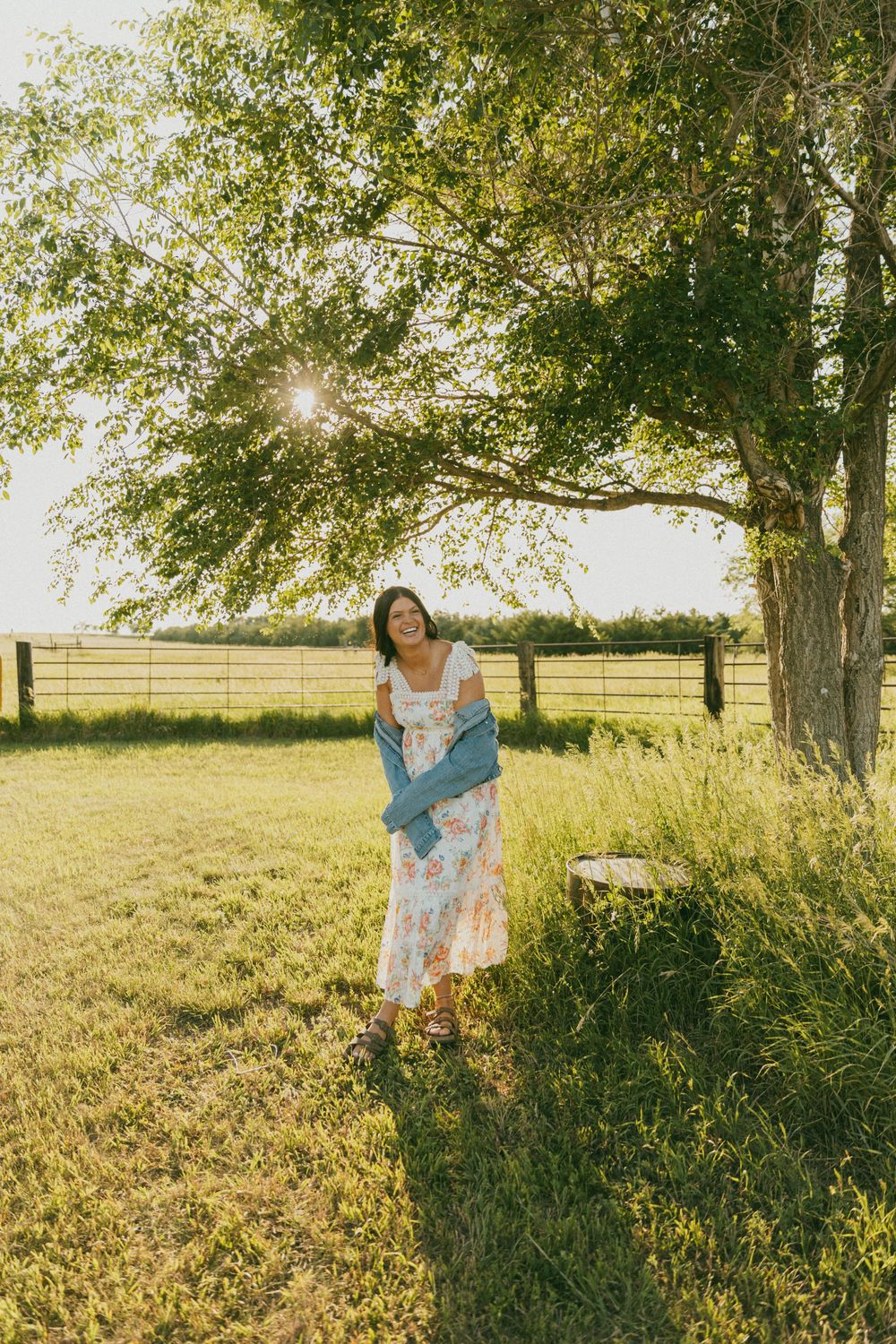 A person in a floral dress stands in a sunlit meadow with tall grass and trees during golden hour.