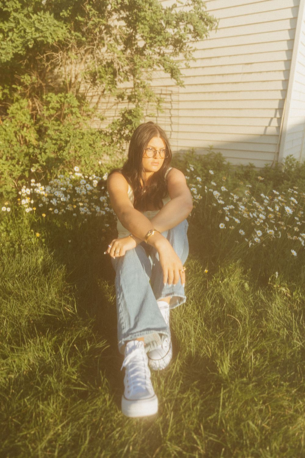 Person in glasses sits on grass wearing jeans and sneakers against white house siding in warm sunlight.