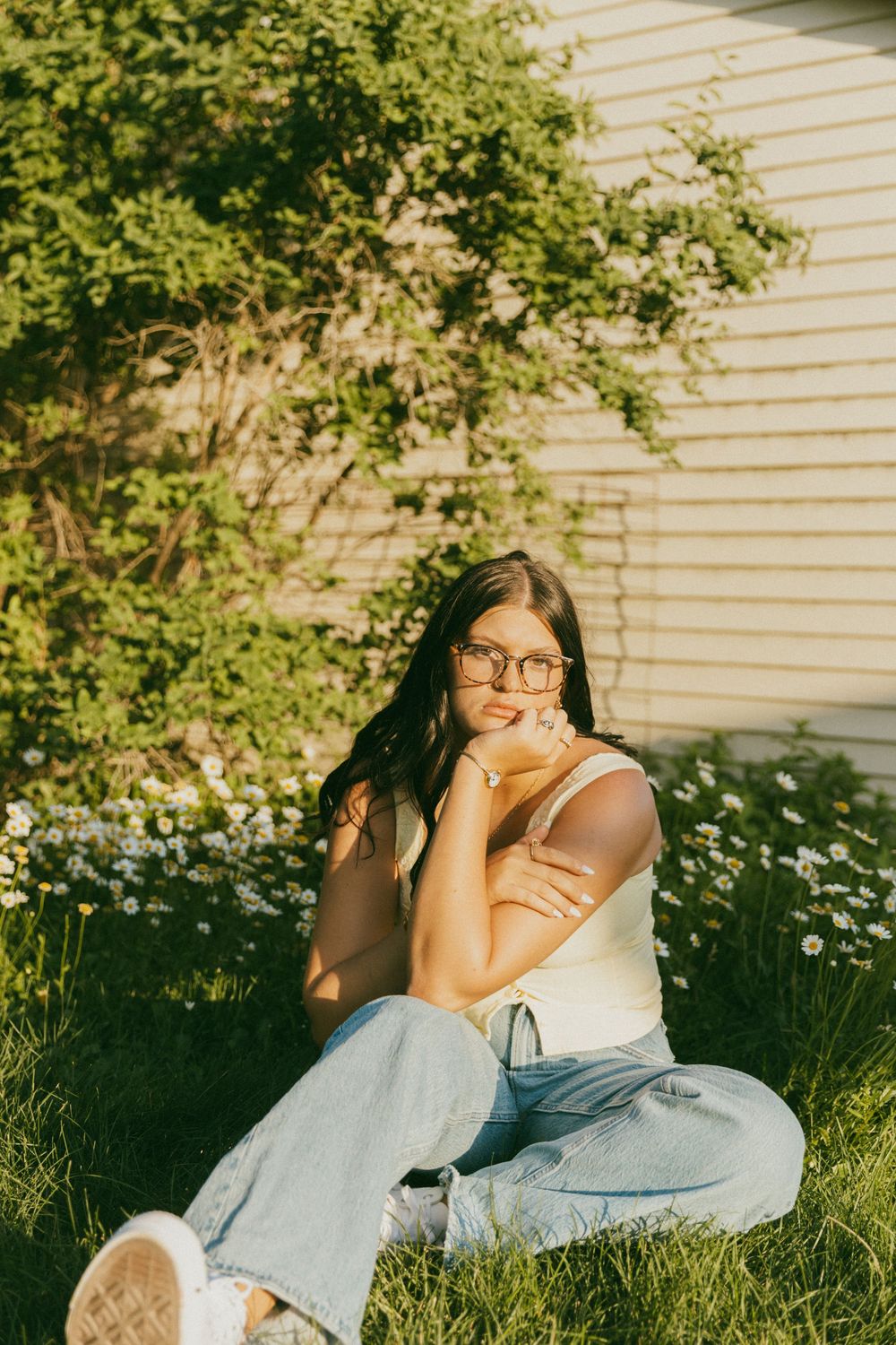 Person in white tank top and jeans sits cross-legged on lawn with flowering bushes and house in background.