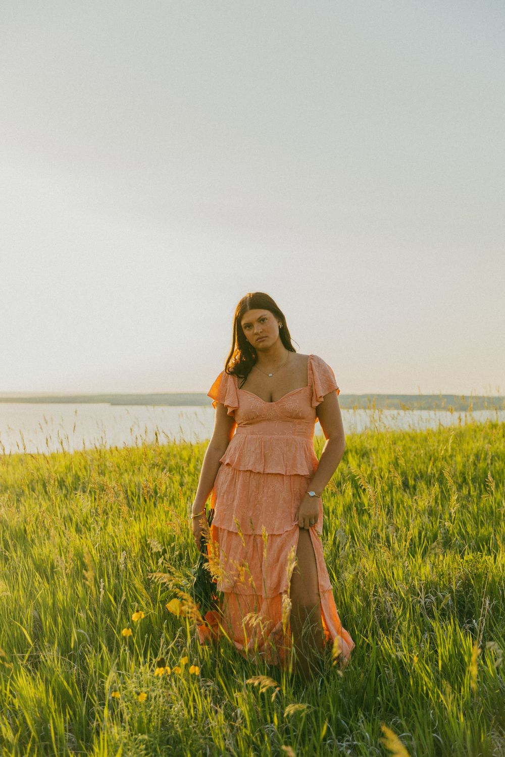 A person in a peach-colored dress stands in a sunlit meadow of tall grass during golden hour.