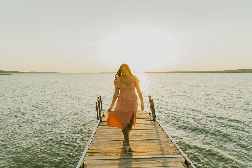A person in an orange dress walks on a wooden dock over calm waters at sunset.