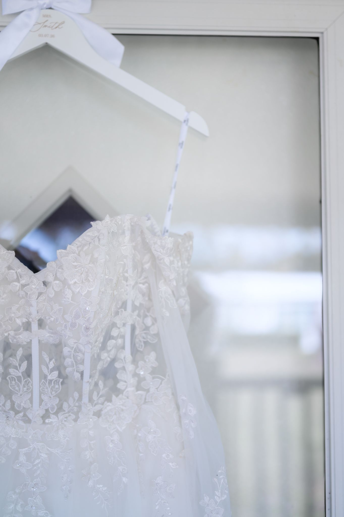 A delicate white lace wedding dress hangs on a white hanger against a light background.