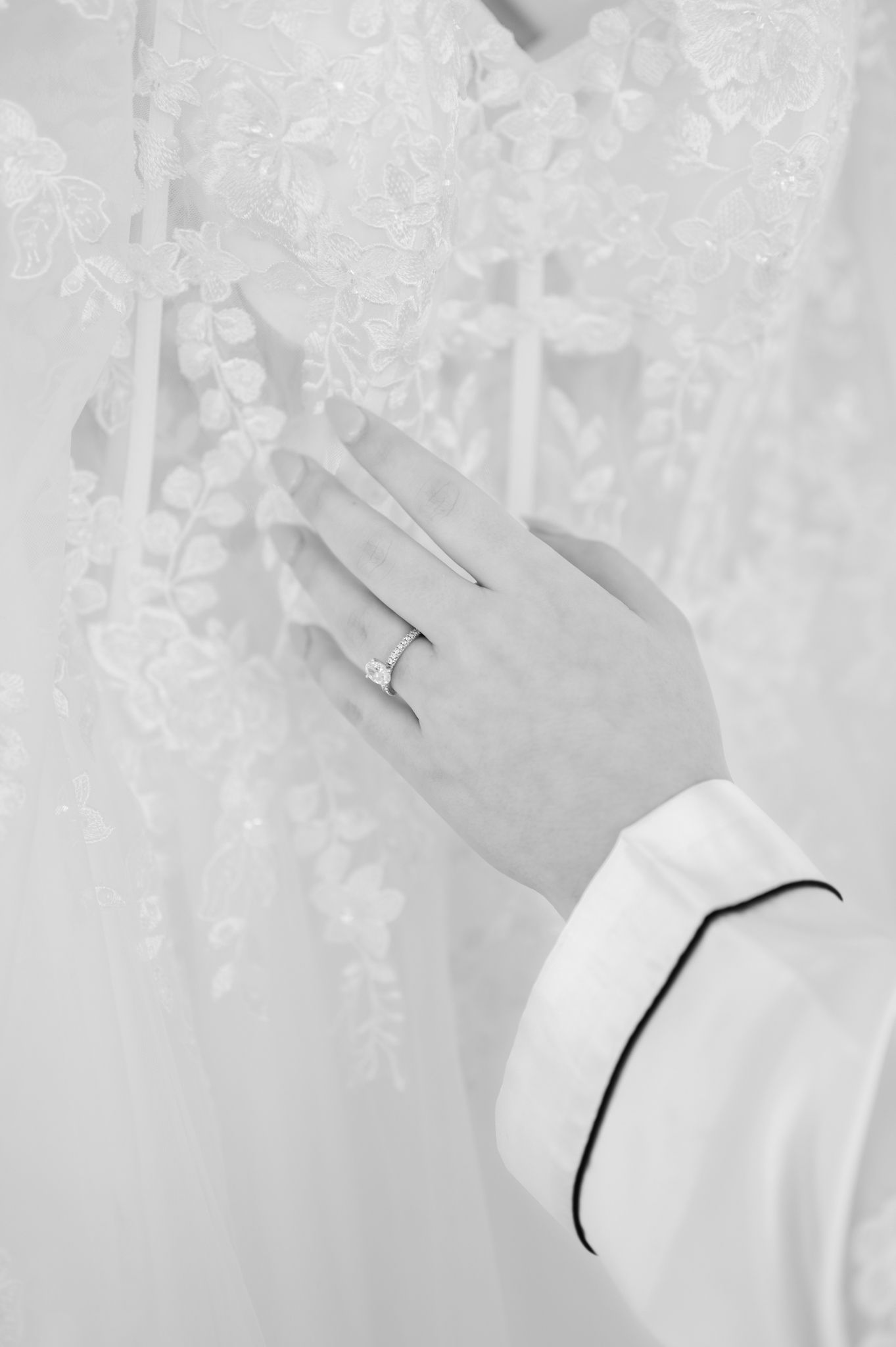 Close-up black and white photo of a hand wearing an engagement ring against delicate lace fabric.