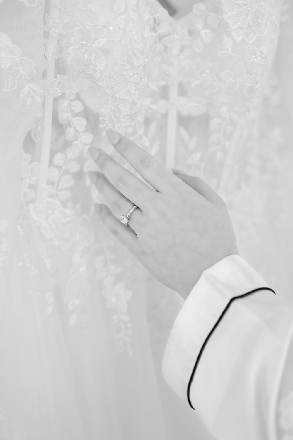 Close-up black and white photo of a hand wearing an engagement ring against delicate lace fabric.