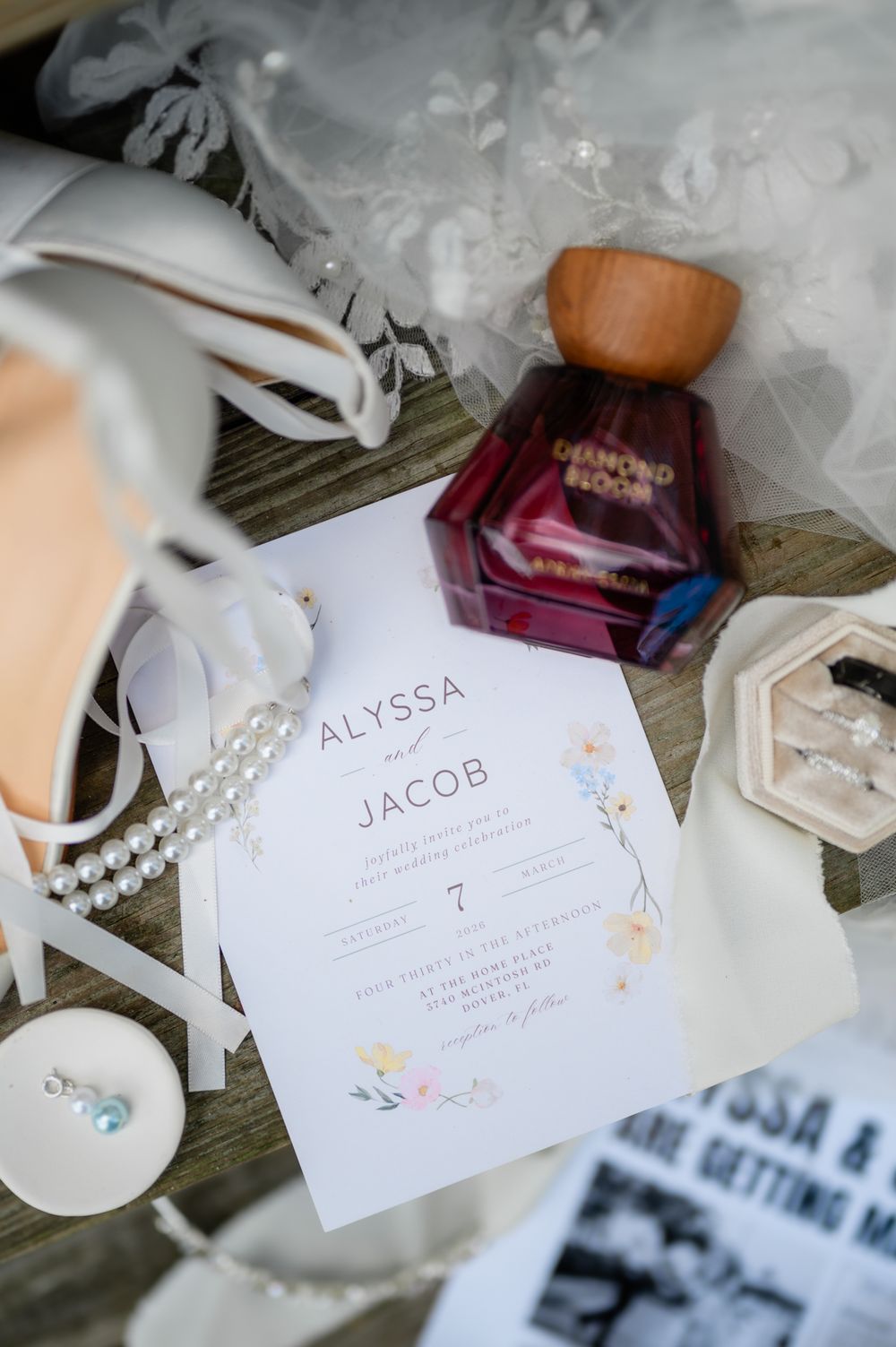 Wedding invitation with names Alyssa and Jacob displayed alongside perfume bottle and pearl accessories.