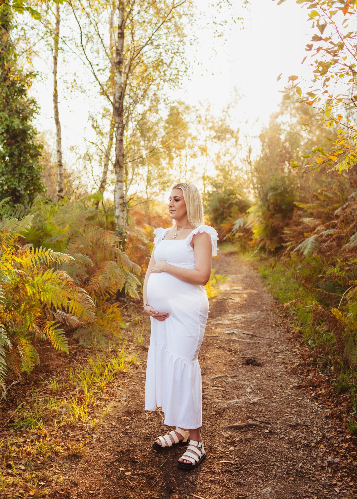 A figure in a white dress stands on a sunlit forest path during golden hour with autumn foliage in the background.