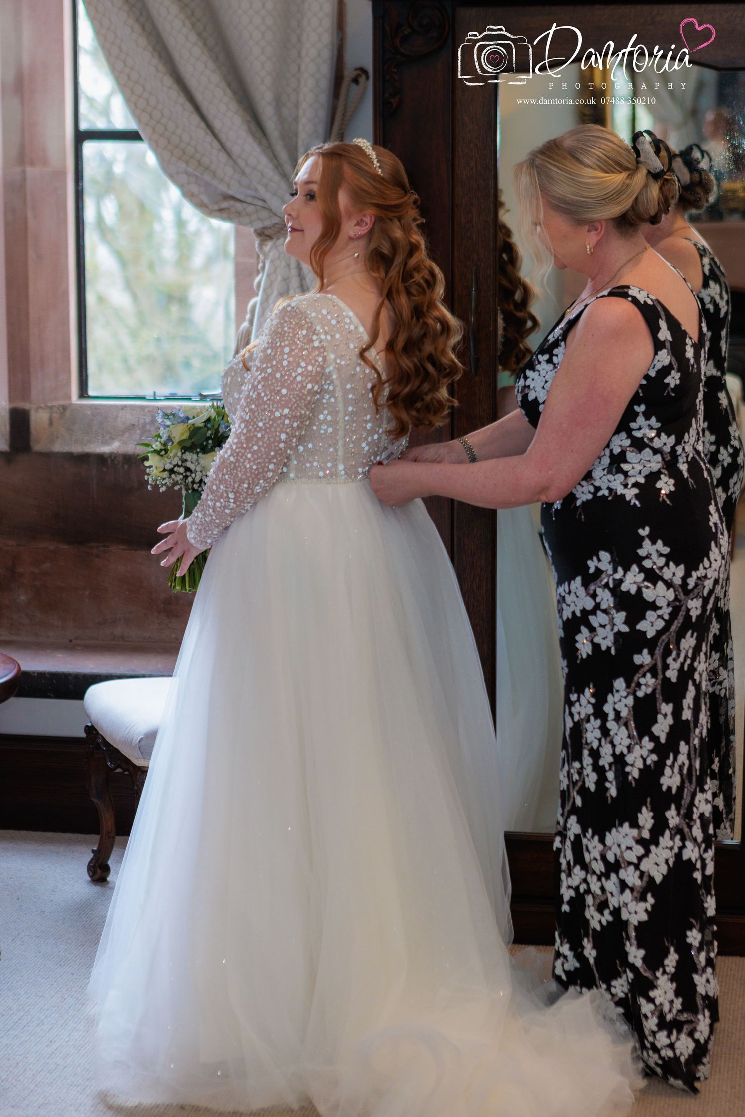 A bride in a white tulle ballgown gets final adjustments to her dress while standing in a rustic venue.