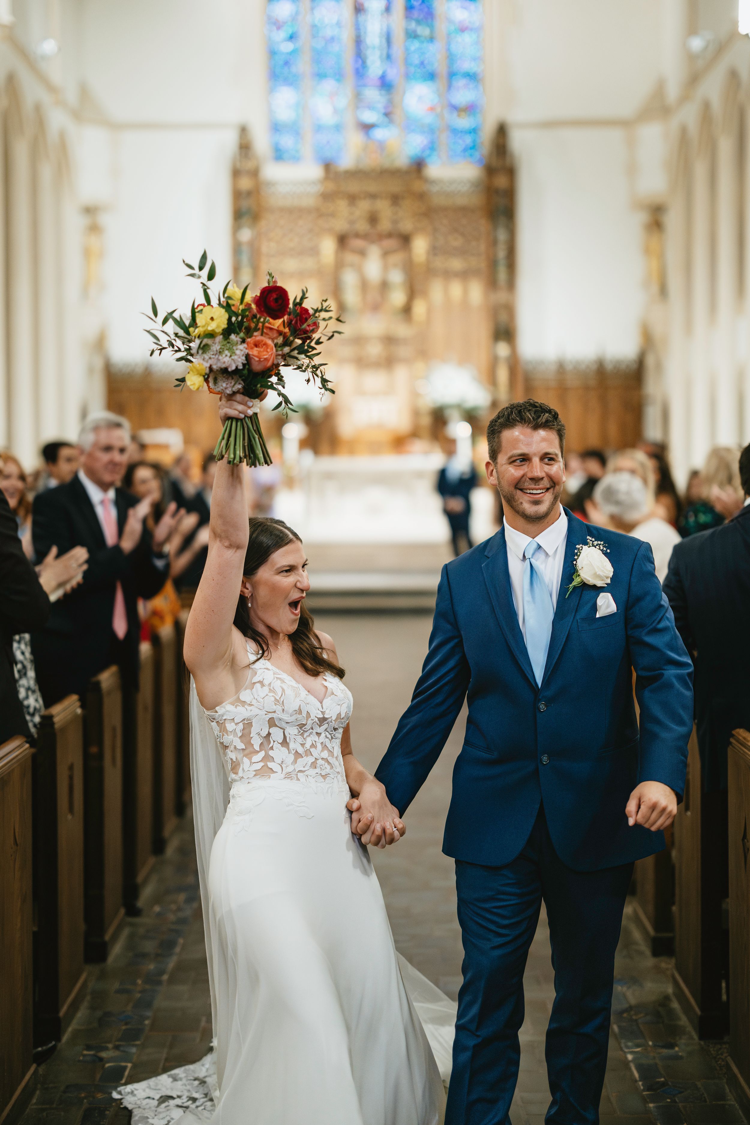 Newlyweds celebrate with joyful raised arms while walking down the church aisle after their wedding ceremony.