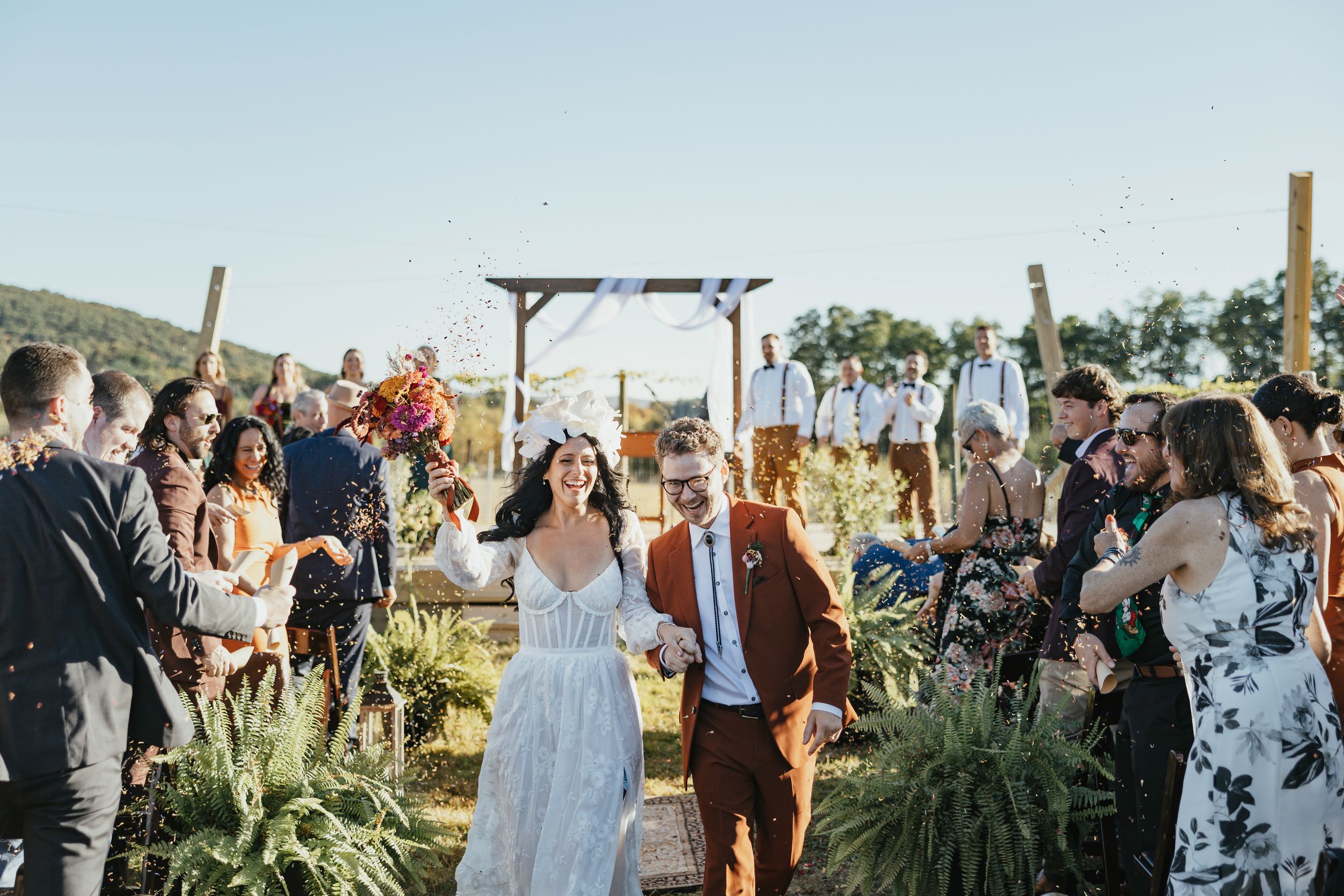 Outdoor wedding ceremony at sunset with guests seated in rows under a wooden pergola in a natural landscape setting.