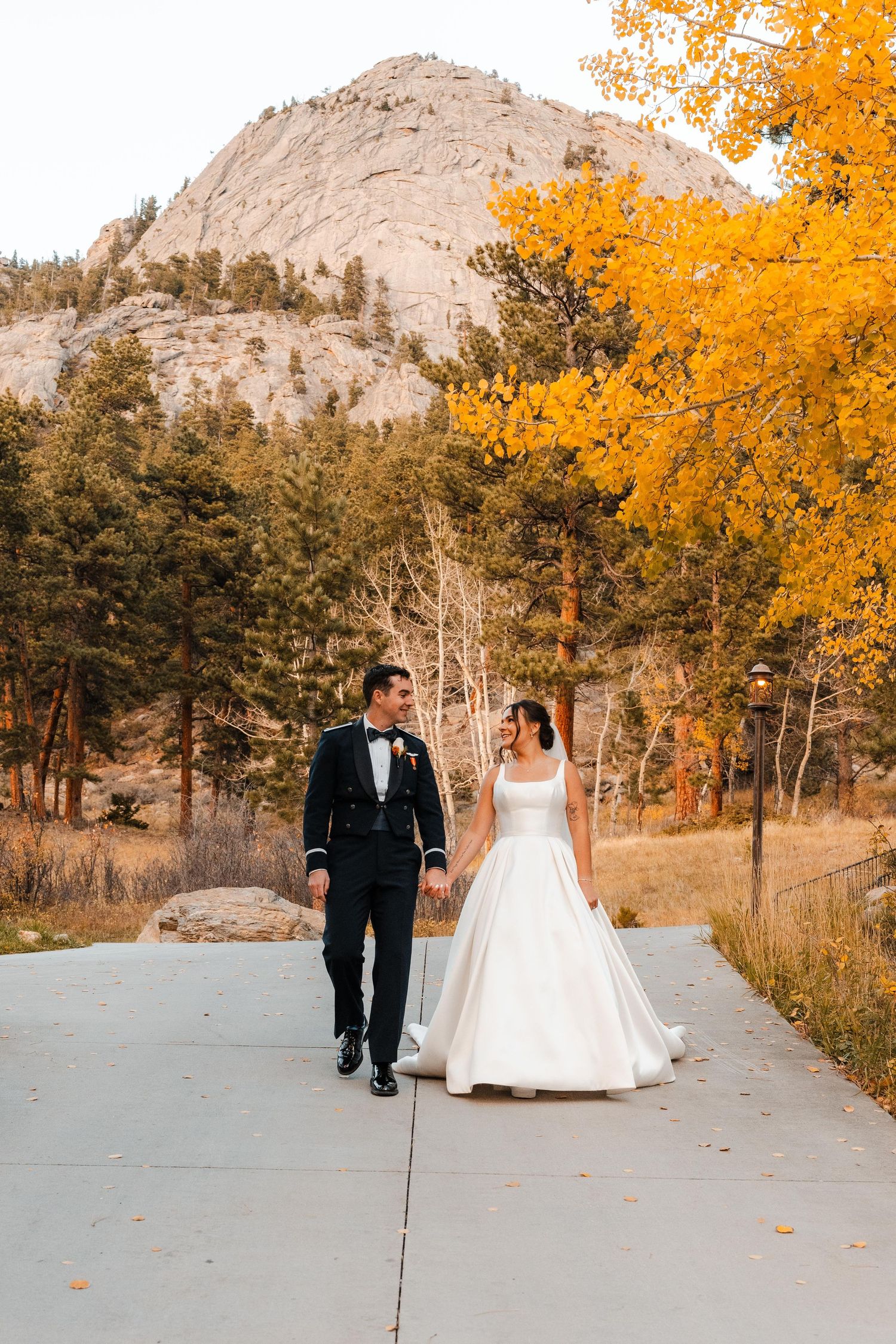 Bride and groom walking hand in hand with golden autumn trees and rocky mountain backdrop.
