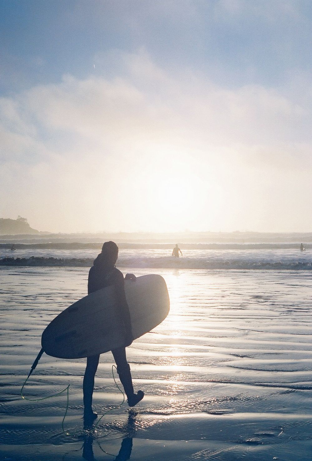Silhouette of a surfer carrying a surfboard on a misty beach at sunset with golden light reflecting on wet sand.