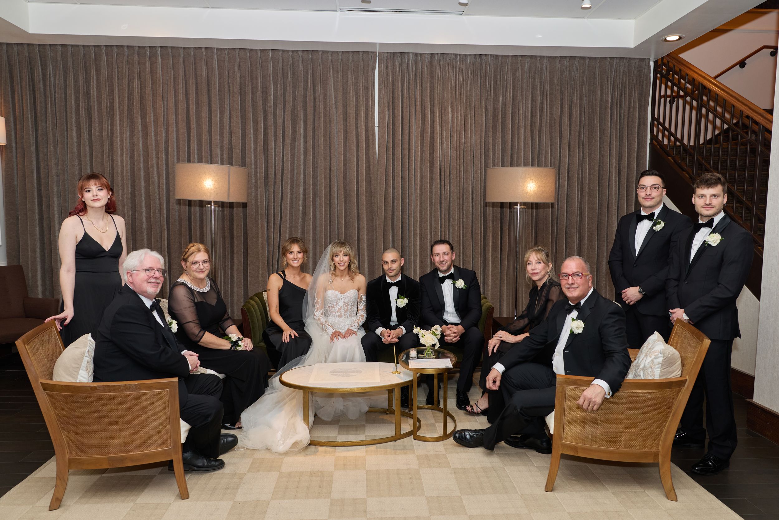 Wedding party in formal attire seated on elegant wooden benches in a modern indoor venue.