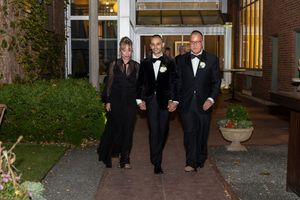 Wedding party members in black tuxedos walk along a brick path outside a building at dusk.