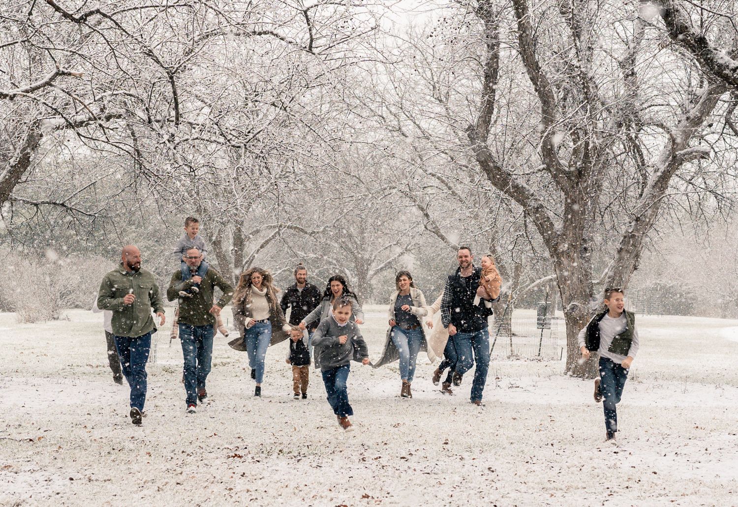 A group of people joyfully running through a snow-covered orchard with bare winter trees overhead.