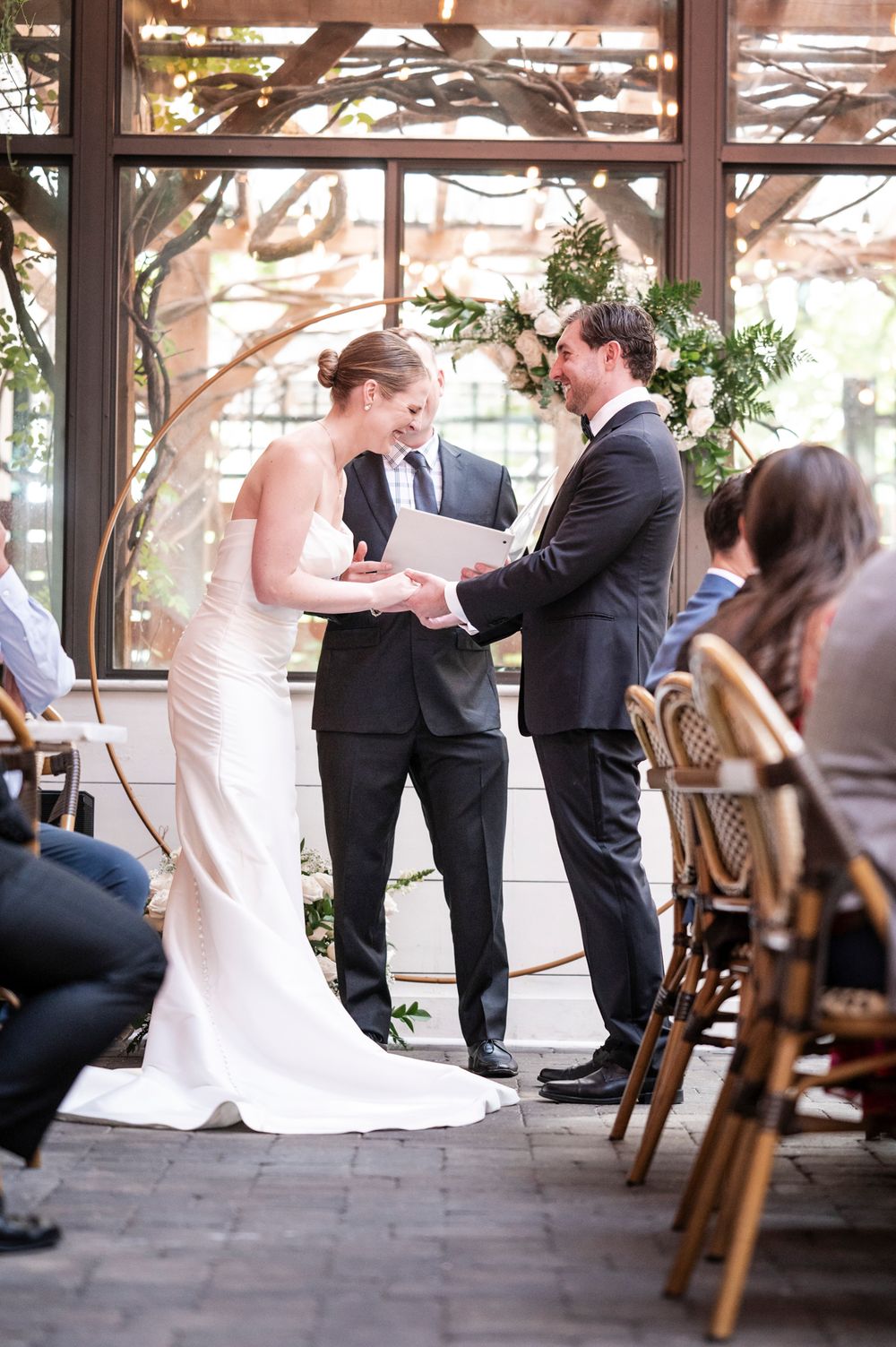 Wedding ceremony taking place in a bright indoor venue with wooden chairs and natural lighting through large windows.