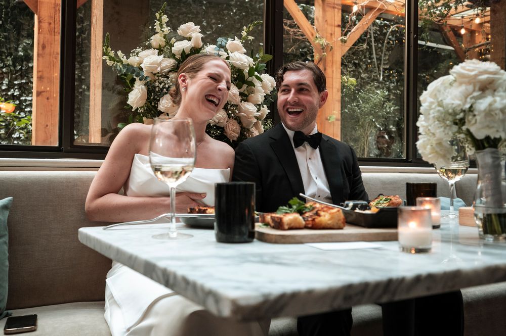 A couple in wedding attire shares a joyful moment while dining at a marble table with floral arrangements.