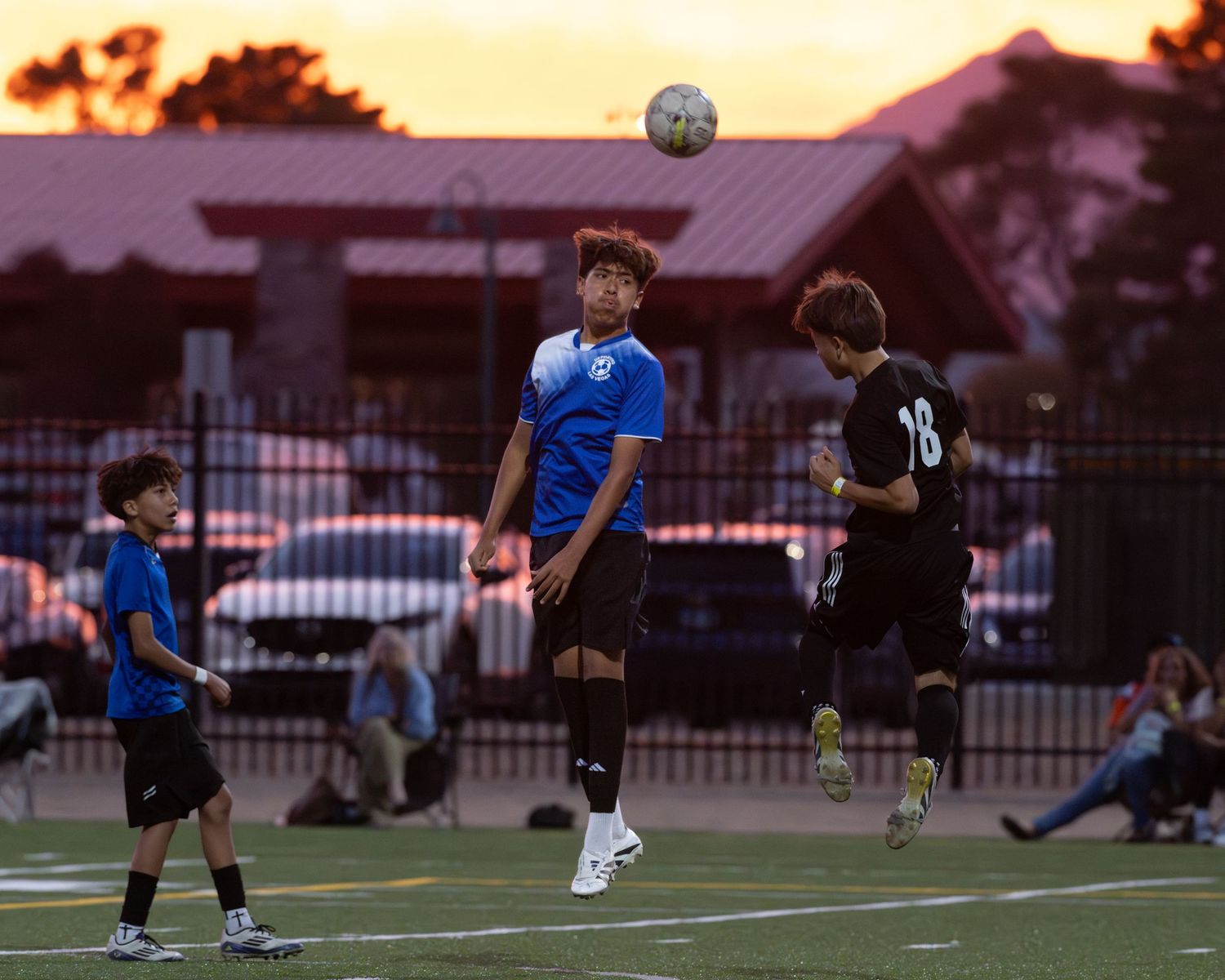 Soccer players compete for a header ball during a sunset match on an outdoor field.