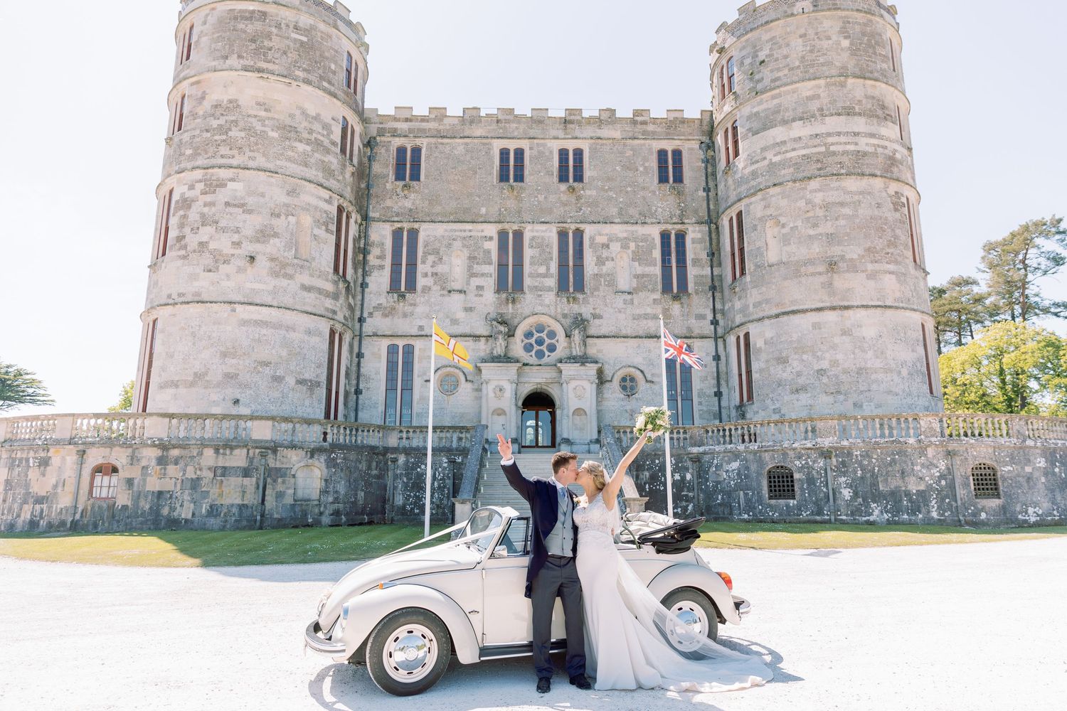 A wedding couple poses with a vintage car outside a grand stone castle on their special day.
