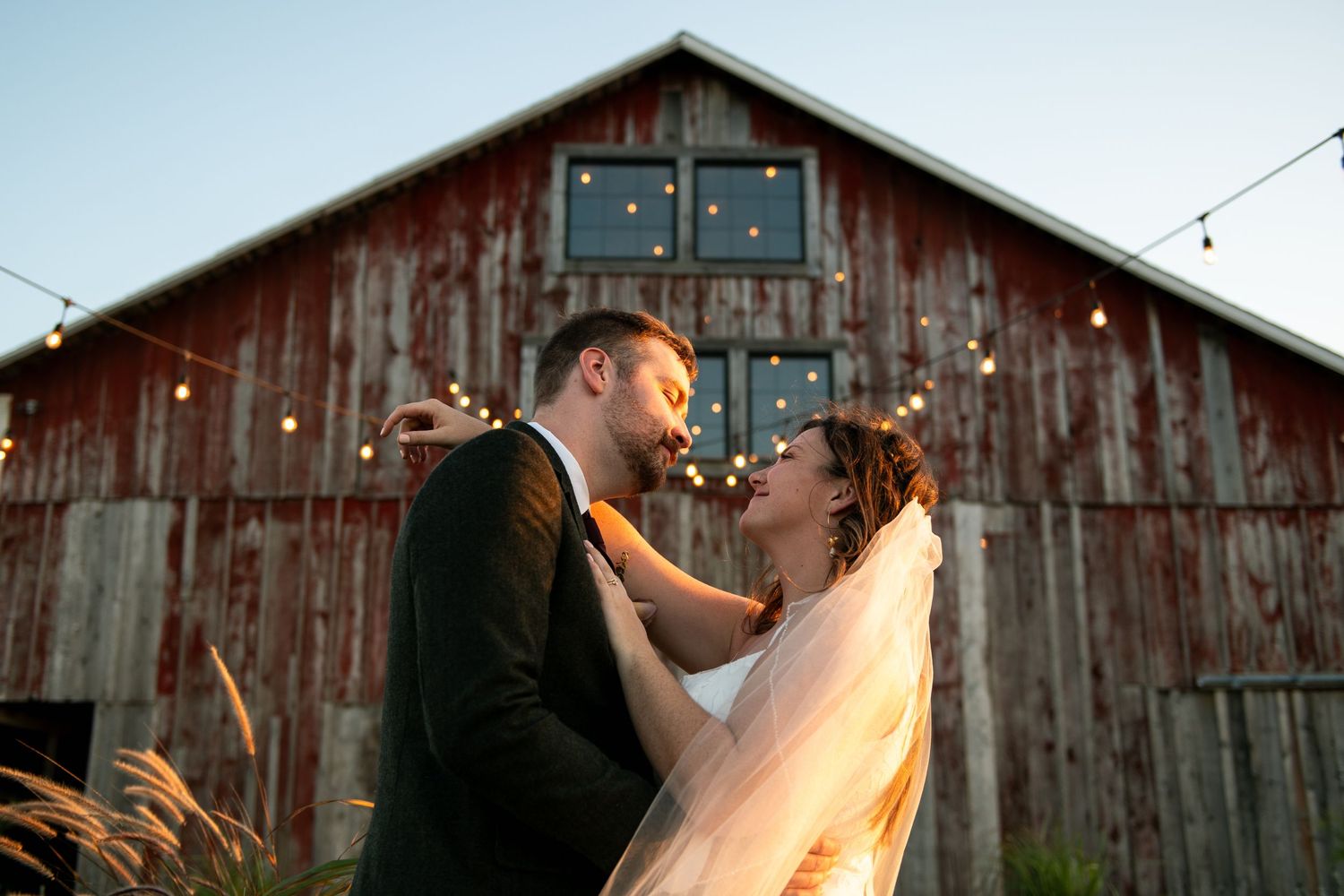 A romantic couple embraces and kisses at sunset in front of rustic wooden barns.