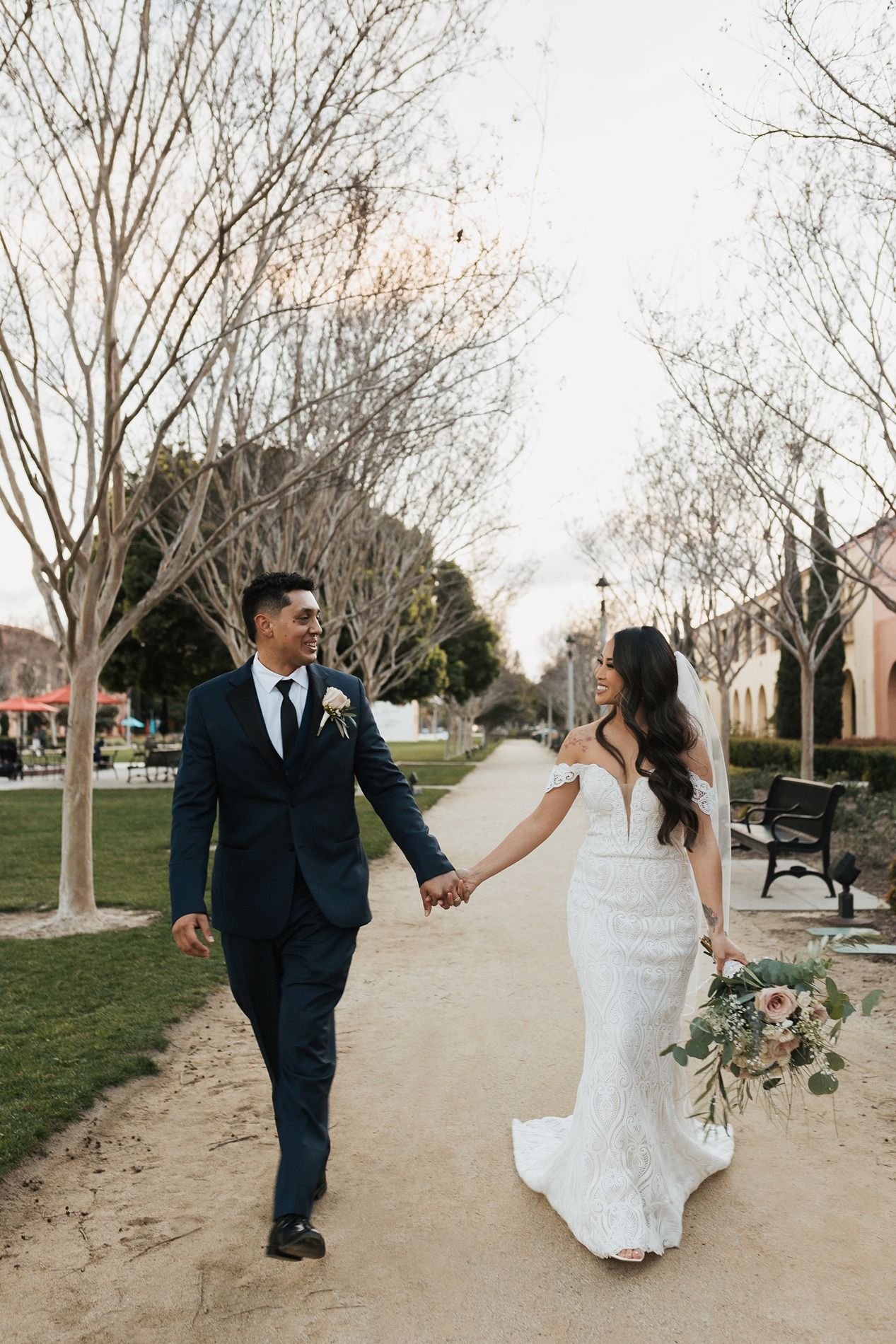 A couple in wedding attire walks hand in hand along a tree-lined path at sunset at Liberty Station in San Diego.