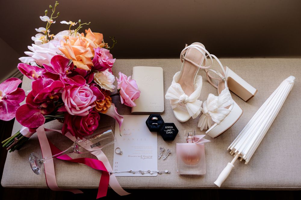 Wedding details flatlay with pink bouquet, white shoes, and stationery on beige surface.