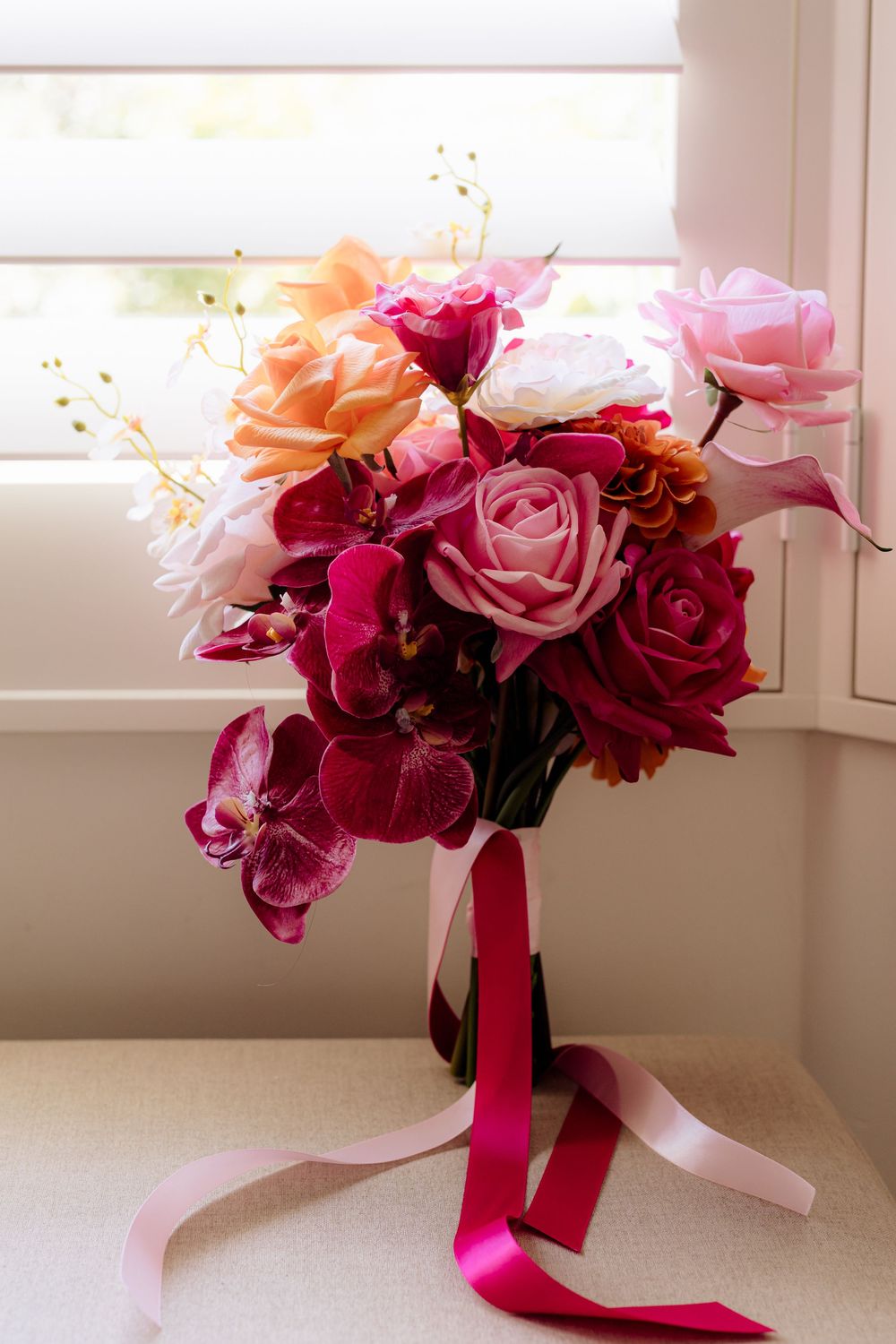 Elegant bridal bouquet with deep red and pink roses tied with flowing ribbons by a window.