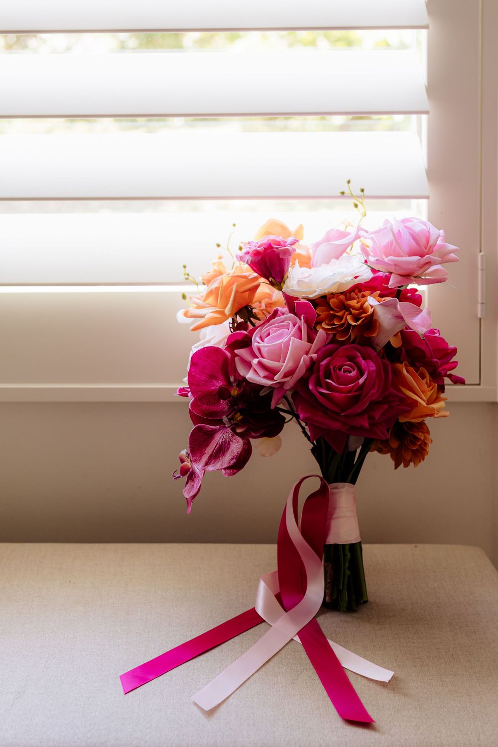 Vibrant pink and orange wedding bouquet with ribbons sits by a window.