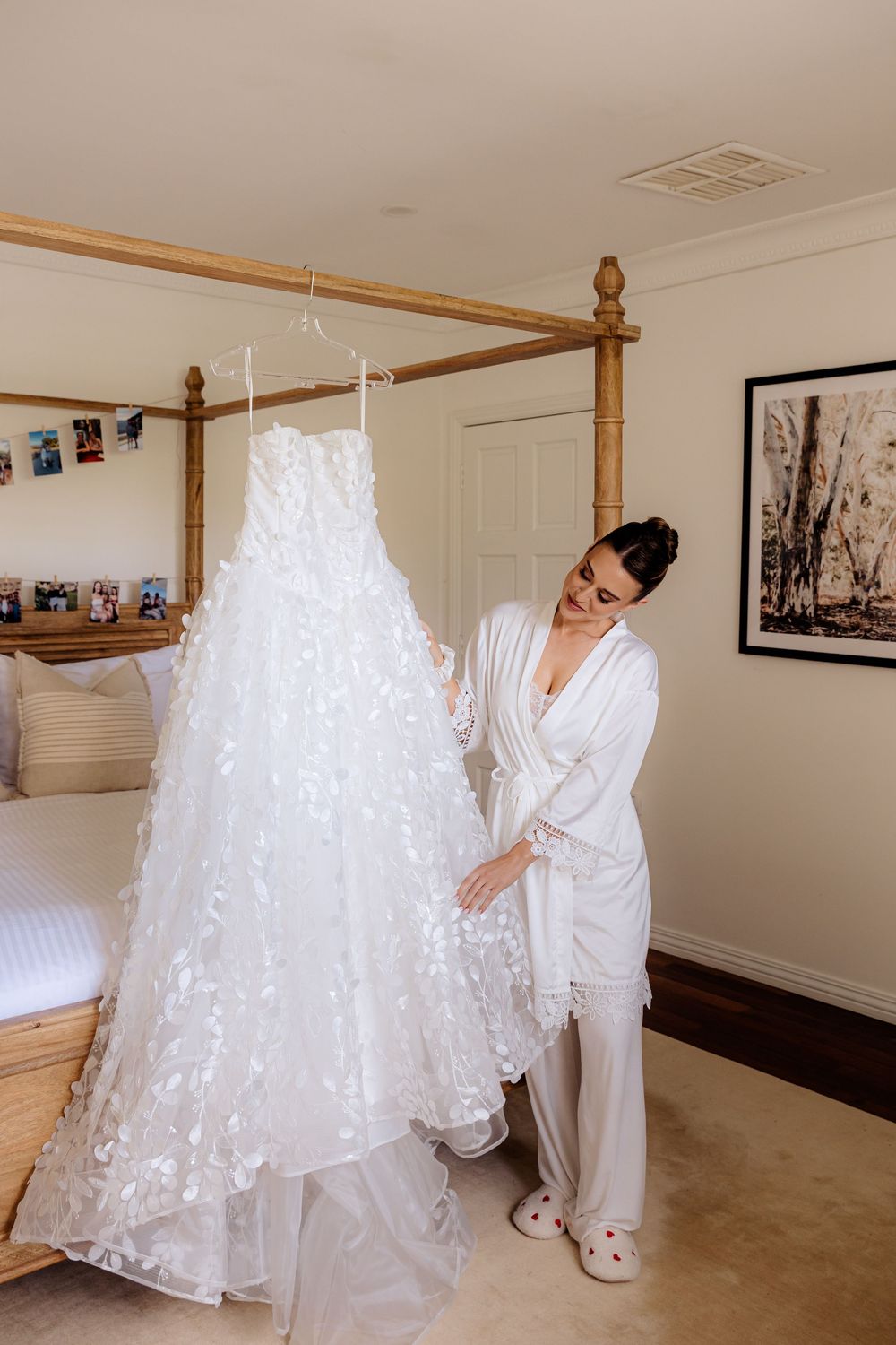 A detailed lace wedding dress hangs from a wooden four-poster bed frame in a bright bedroom.