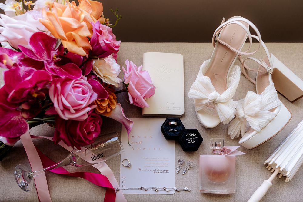 Wedding details flatlay with pink roses, white heels, perfume bottle and invitation on light background.