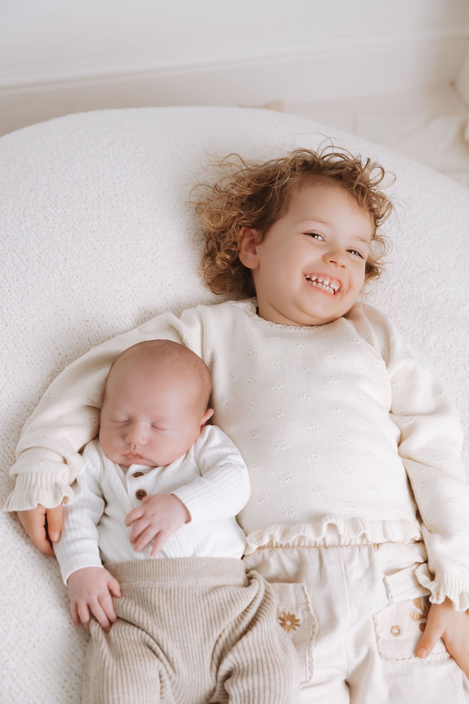 Two children in white outfits lay together on a white surface, one smiling while the other sleeps peacefully.