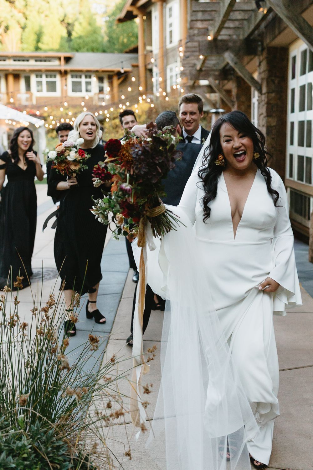 Wedding party walking and laughing together on outdoor patio with autumn decor and string lights overhead.