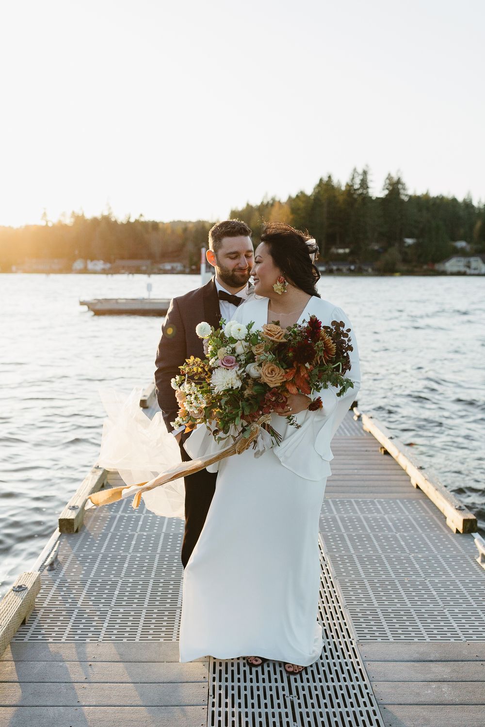 Bride and groom share a romantic moment on a lakeside dock at golden hour, bride holding a lush autumn bouquet.