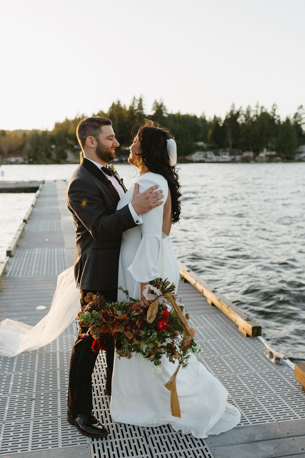 Bride and groom embrace on a lakeside dock at sunset, she holds a lush fall bouquet in a white gown.