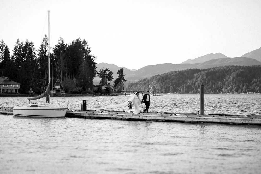 Black and white photo of a couple dancing on a dock by a lake with sailboat and mountains in background.