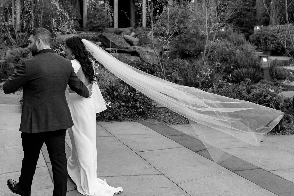 Black and white photo of bride and groom walking, her long veil flowing in the wind at an outdoor wedding.