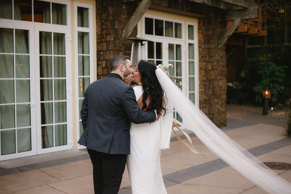 Bride and groom share a romantic first kiss outdoors, with brides veil flowing, in front of an elegant stone building.