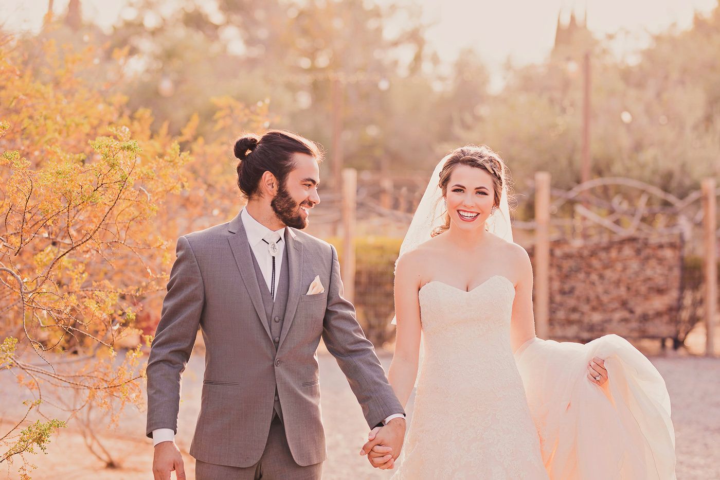 A couple in formal wedding attire walks together through a lush garden path surrounded by trees.