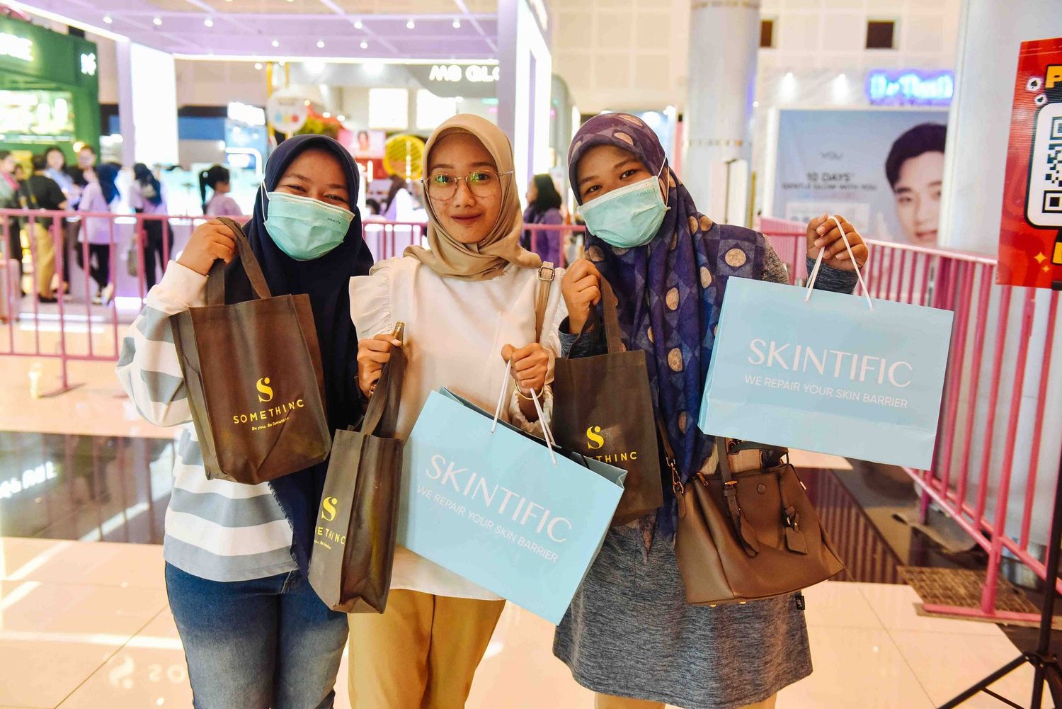 Three shoppers wearing hijabs and face masks hold shopping bags and display signs at a retail mall.