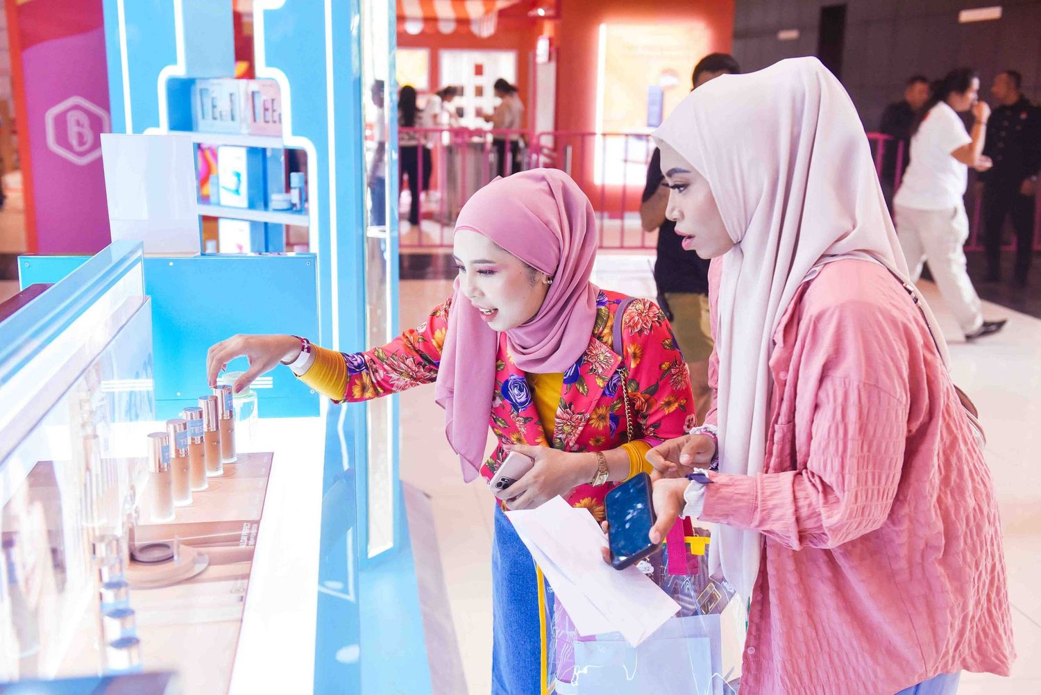 Two women wearing hijabs examine products at a bright blue retail display counter in a shopping center.