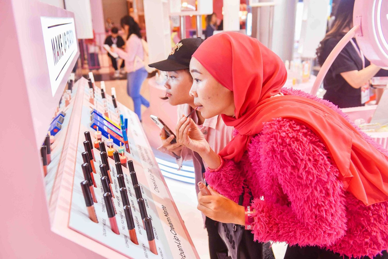 Women in bright pink and red clothing try on makeup products at a beauty counter display.