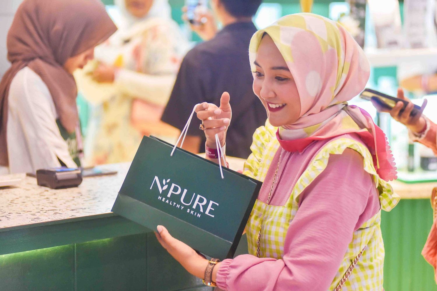 Shopper in pink hijab smiles while holding a green N'PURE shopping bag at a retail store.