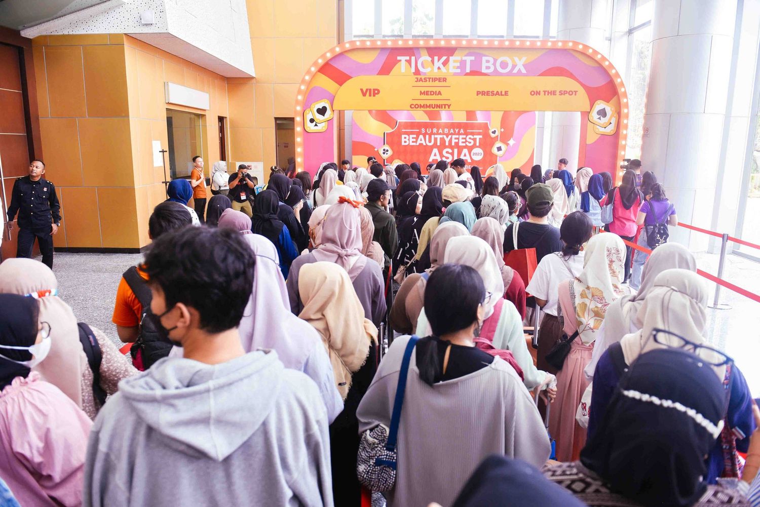 A crowded indoor event with people gathered in front of a red and orange decorated stage or backdrop.