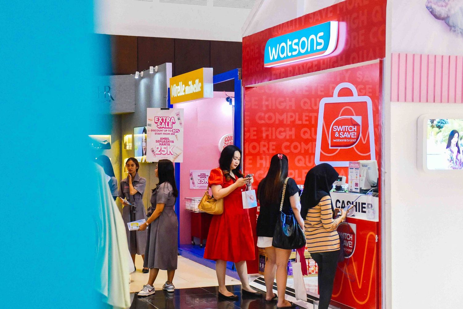 Shoppers browse colorful retail displays in a modern mall with bright turquoise walls and red signage.