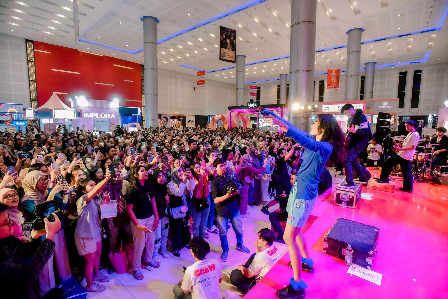 A large crowd of people gathers around a brightly lit stage area with pink and purple lighting at an indoor event.