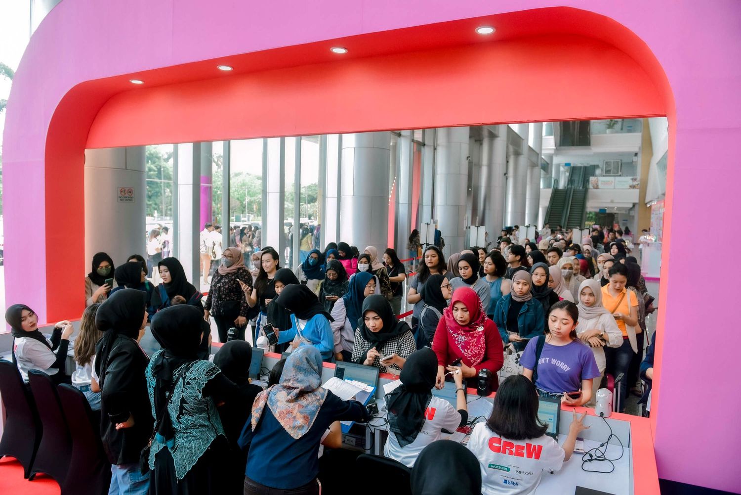 A crowded line of people waiting under a pink archway in what appears to be a shopping mall or event space.