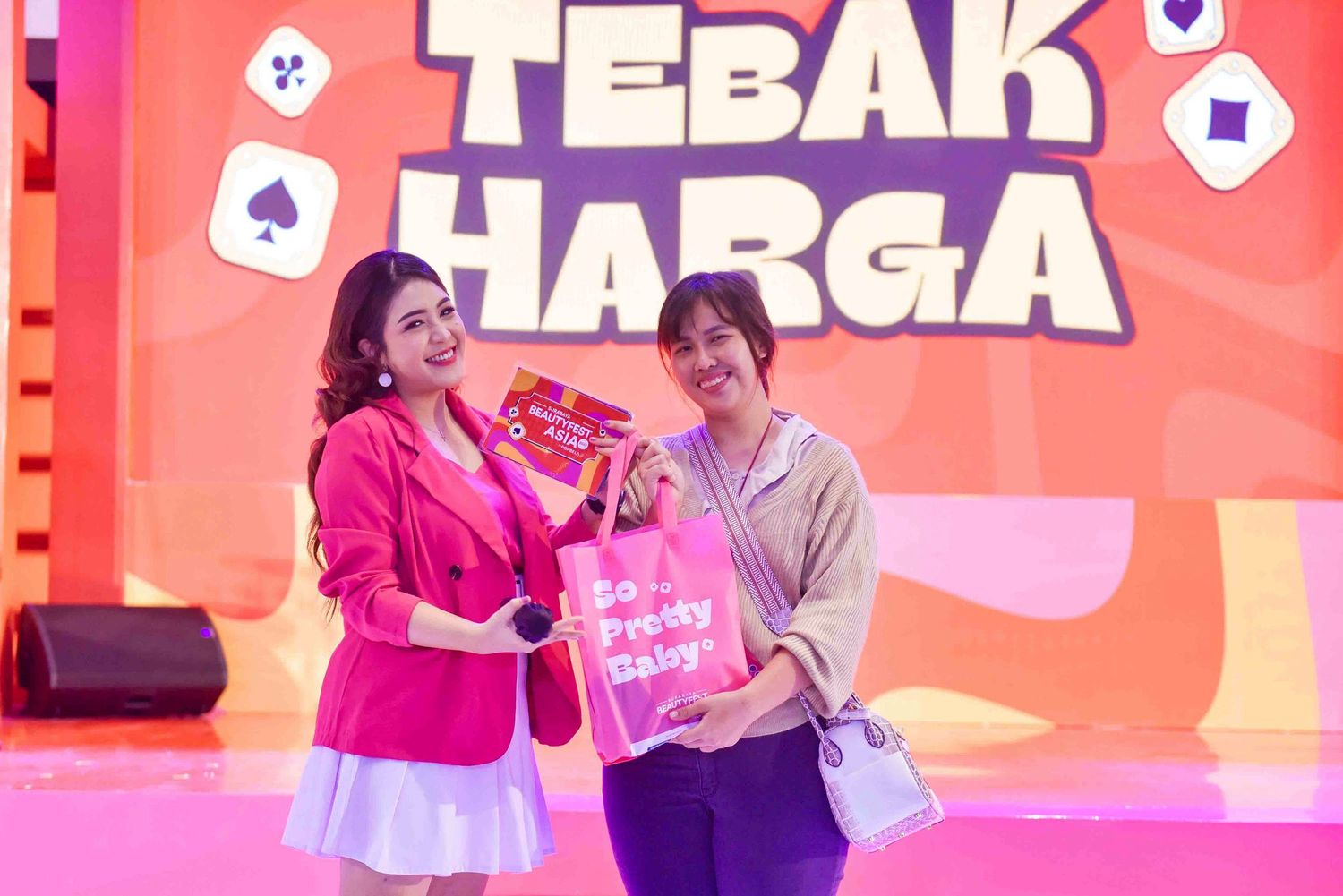 A group of people playing a game show on a brightly lit pink stage with playing card symbols in the background.