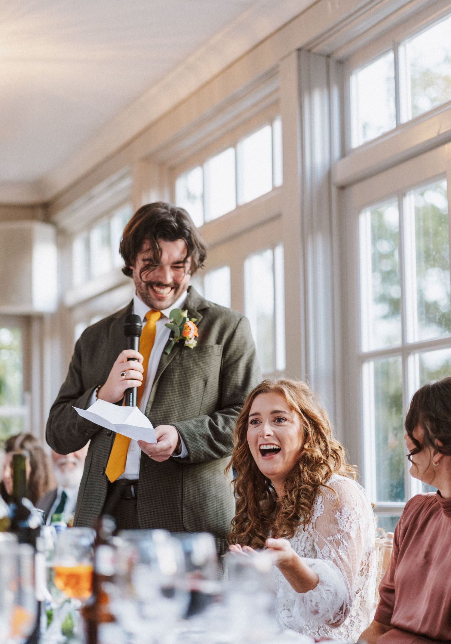 Wedding speech moment captured during reception with laughter and joy in a bright sunlit venue with large windows.